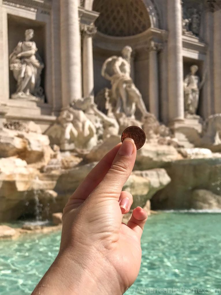 hand holding coin in front of Trevi Fountain